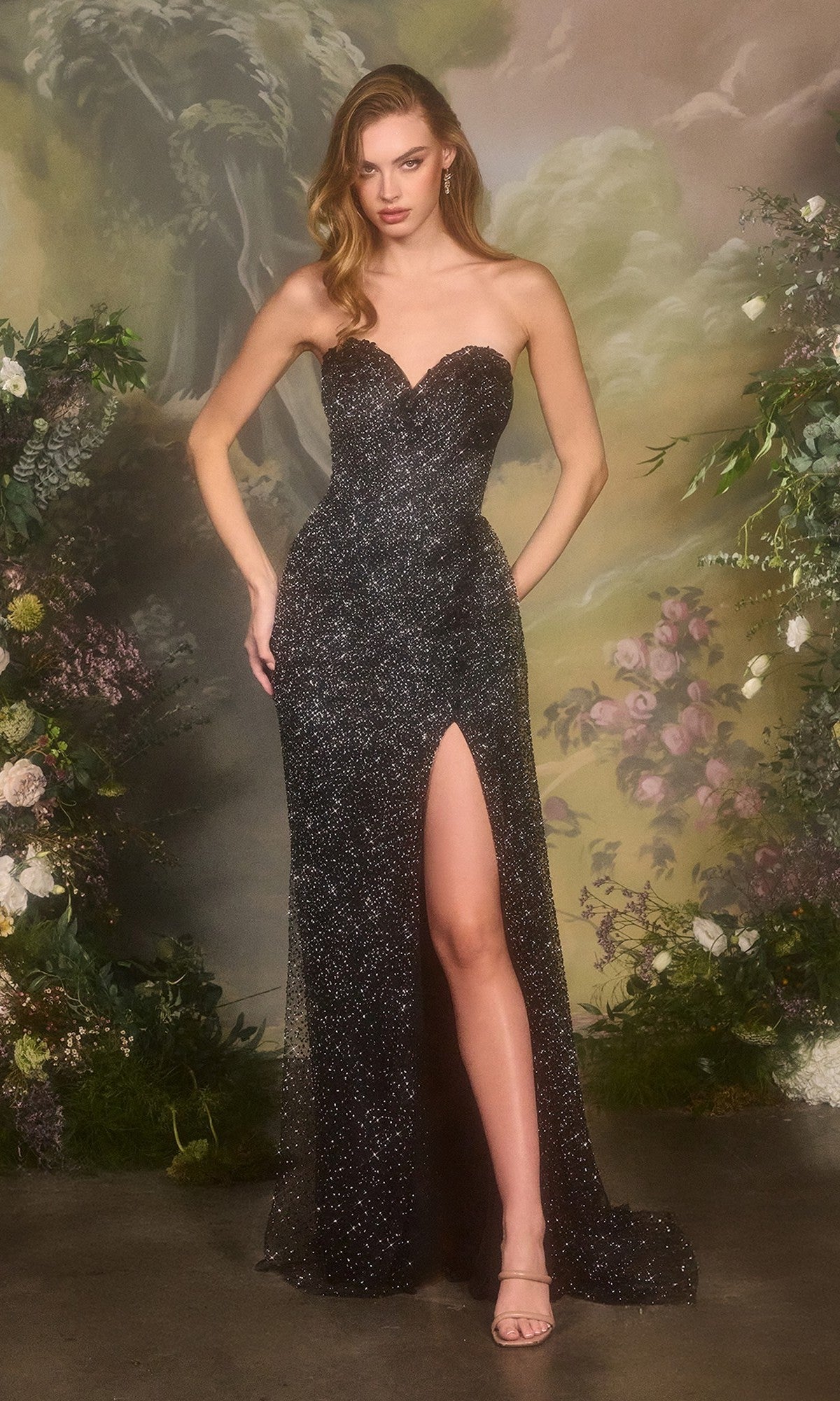 A woman poses before a floral backdrop in the Andrea and Leo A1494 long black prom dress, featuring a strapless beaded corset bodice, floor-length glittery skirt with high slit, and nude heels.